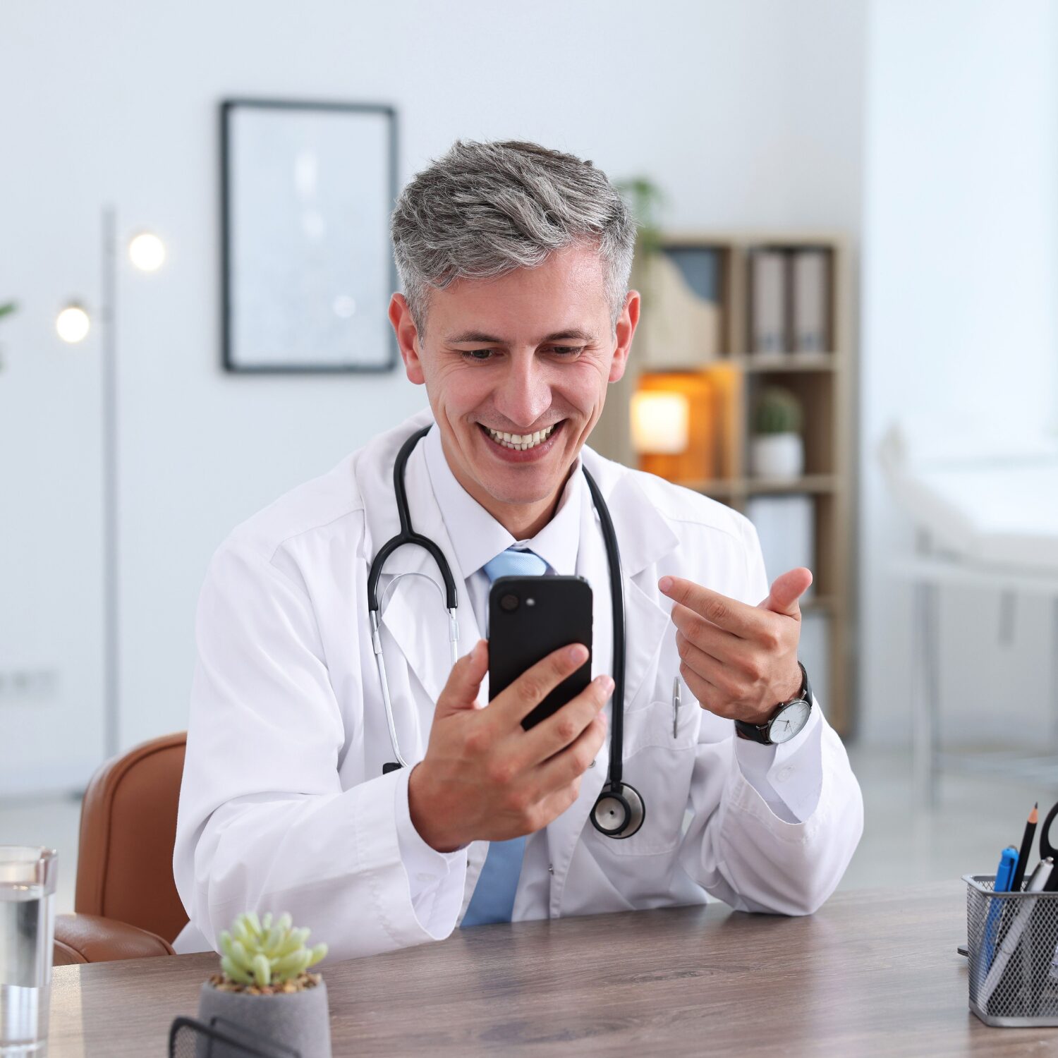 Doctor smiling while using smartphone during telemedicine consultation at medical office desk